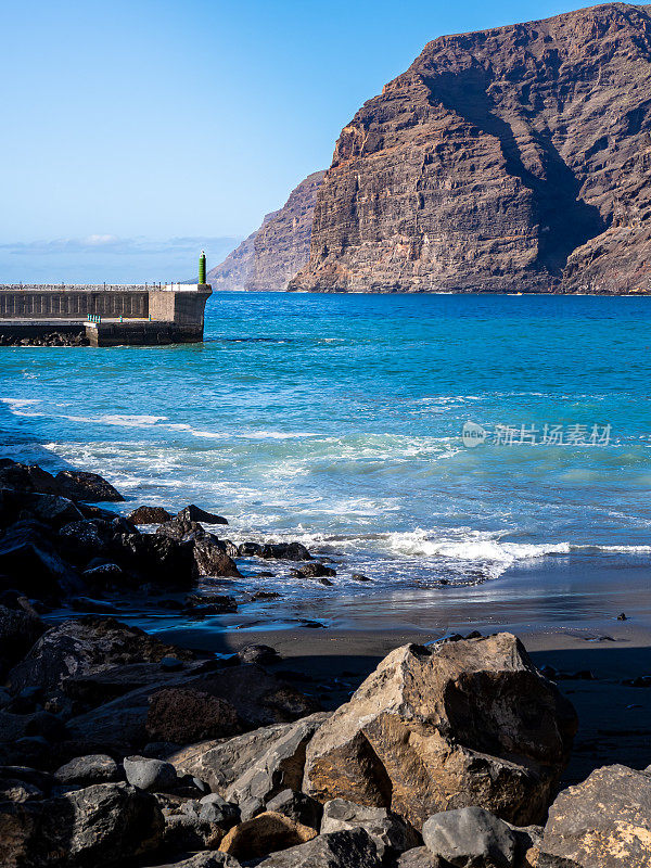 Acantilados de Los Gigantes cliffs provide a backdrop to the Playa Los Guíos black beach with the Los Gigantes harbor entrance and rugged rocks in the foreground, creating a scenic travel destination.